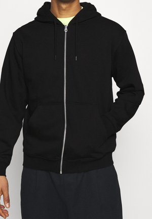 Hombre vistiendo una sudadera negra con cremallera, bolsillos delanteros y capucha ajustable con cordón, combinada con pantalones oscuros y una camiseta interior de color claro.