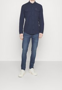Camisa de botones azul marino con dos bolsillos en el pecho, combinada con jeans ajustados azules y zapatillas blancas, presentada sobre un fondo neutro.
