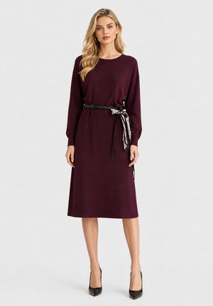 Donna che indossa un vestito lungo fino al ginocchio, a maniche lunghe, viola scuro, con una cintura nera legata con una sciarpa fantasia, abbinato a décolleté neri.