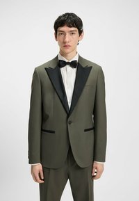 Olivgrüner Smoking-Jackett mit schwarzen Satin-Revers, Ein-Knopf-Verschluss und zwei aufgesetzten Taschen, kombiniert mit einem weißen Hemd und einer schwarzen Fliege.