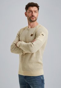 Jasne, beżowe sweter dziany z teksturowanym wzorem, ściągaczami przy mankietach i dolnej krawędzi oraz małym logo na klatce piersiowej. Noszony z niebieskimi dżinsami.