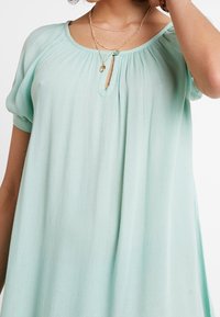 Blouse vert menthe clair en tissu doux et plissé. Présente un col rond avec un détail en trou de serrure et des manches courtes et bouffantes.