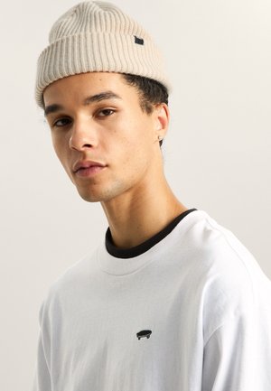 Beige ribbattu beanie, joka on yhdistetty valkoiseen crewneck-paitaan, jossa on pieni musta skeittigrafiikka rinnassa ja musta kaulus.