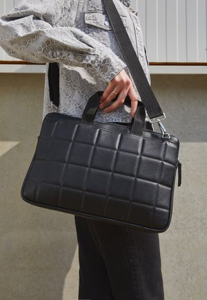 Persona vistiendo una camisa gris con estampado y pantalones negros, sosteniendo un bolso de cuero negro acolchado con asas y correa para el hombro.