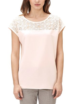 Femme portant un chemisier rose clair à manches courtes avec des détails en dentelle sur le haut de la poitrine et un pantalon gris à motif, debout devant un fond blanc.