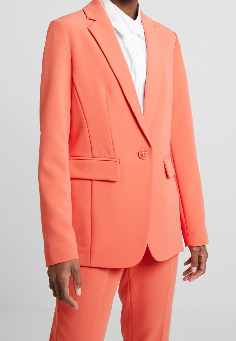 Koralowo-pomarańczowa marynarka z gładkiego materiału, z pojedynczym zapięciem na guzik, klapami z wycięciem i bocznymi kieszeniami. Widoczne spodnie w zestawie.