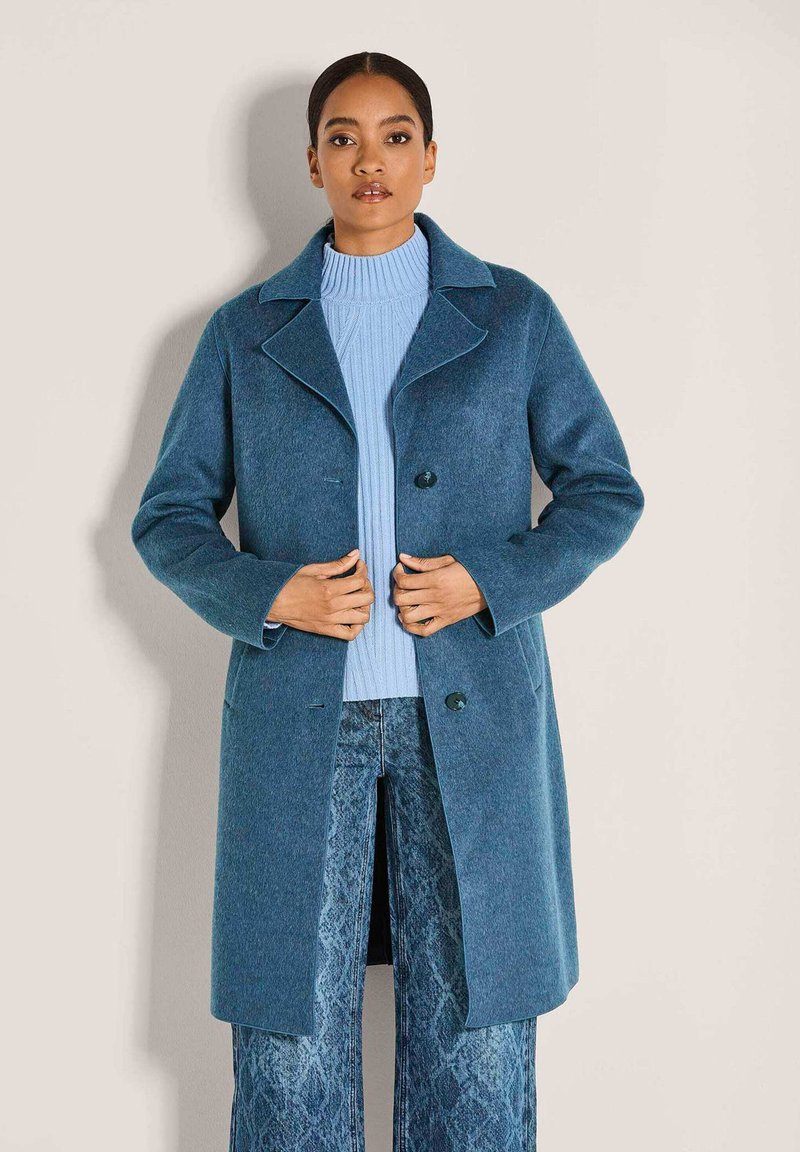 Vrouw gekleed in een lange blauwe jas over een lichtblauwe coltrui en gestreepte blauwe wijde broek, staand tegen een effen achtergrond.