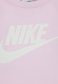 Jasnoróżowa bluza wykonana z teksturowanej tkaniny, z dużym białym logo "NIKE" i charakterystycznym znakiem Swoosh umieszczonym na przodzie.
