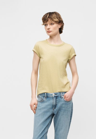 Νεαρός με κοντά σγουρά μαλλιά, φορώντας ανοιχτοκίτρινη κοντομάνικη μπλούζα με στραμμένα μανίκια και ανοιχτόχρωμο τζιν, στέκεται με το ένα χέρι στην τσέπη.