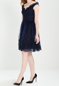 Navy blue dress with lace detailing, off-the-shoulder design, sheer skirt overlay, and scalloped hem, paired with black high heels.