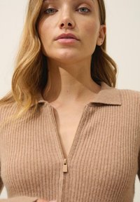 Beige gerippter Strickpullover mit Reißverschlusskragen. Weiche Textur mit tailliertem Design. Reißverschluss in passendem Farbton, minimalistische Details.