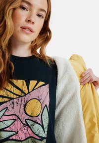 Zwarte t-shirt met kleurrijke grafische print van een zon en bladeren. Model houdt een gele buitenjas vast met een witte, pluizige voering.