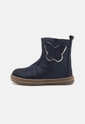 Bottine en suède bleu marine avec une application de papillon pailleté, bout rond, panneau latéral élastique et semelle en caoutchouc texturé.