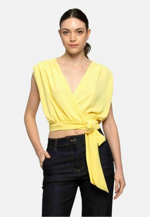 Top giallo avvolgente con maniche corte e scollatura a V, annodato in vita. Tessuto liscio, abbinato a pantaloni in denim scuro, con dettagli minimi.