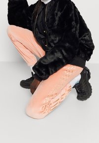 Černá bunda z umělé kožešiny typu bomber, růžové sametové tepláky se svislými pruhy, černé tenisky, v podřepu na jednobarevném pozadí.