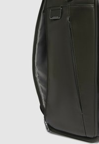 Zaino in pelle verde oliva con trama, dotato di un ampio scomparto, chiusura con zip e una tasca laterale aggiuntiva. Dettaglio di cuciture rinforzate visibile.