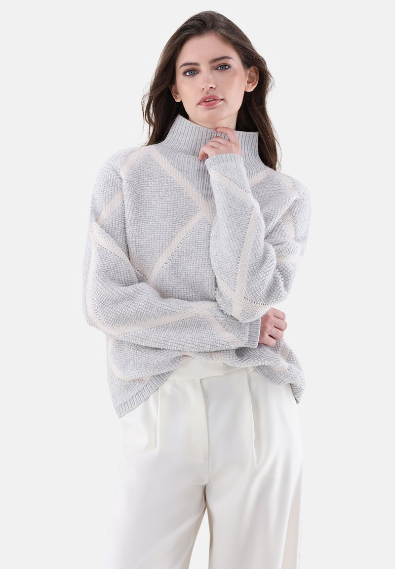 Grauer Strickpullover mit hohem Kragen, der eine strukturierte Oberfläche und diagonale cremefarbene Streifen aufweist. Kombiniert mit cremefarbenen Hosen.