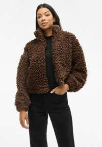 Smeđa polar jakna na patentni zatvarač s teksturiranim, plišanom vanjskom površinom i visokim ovratnikom; nosi se preko crnog topa, u kombinaciji s crnim hlačama.