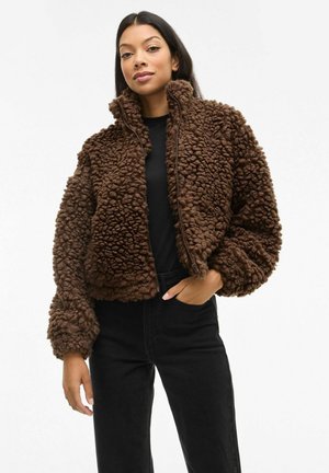 Chaqueta de felpa marrón con cierre, exterior texturizado y peludo, y cuello alto; usada sobre una blusa negra, combinada con pantalones negros.