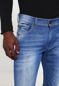 Vaqueros de mezclilla azul con efecto desgastado, ajuste ceñido, diseño de cinco bolsillos y herrajes metálicos. La tela muestra una textura ligera y detalles sutiles de desgaste tipo 'whiskers'.