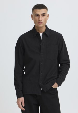 Junger Mann mit kurzen Haaren, der ein schwarzes Hemd mit Knöpfen und schwarze Hosen trägt, steht mit einer Hand in der Tasche vor weißem Hintergrund.