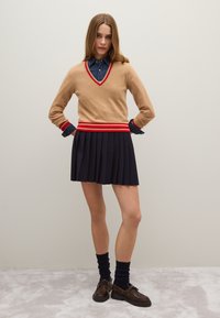 Jersey de color beige con escote en forma de V y rayas rojas y blancas en la cintura, combinado con una falda azul marino con pliegues. Medias oscuras y zapatos marrones completan el conjunto.