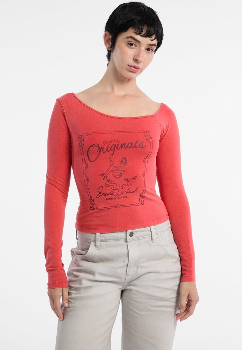 Rotes Langarmoberteil mit weitem Halsausschnitt, das mit schwarzem Drucktext und Grafiken verziert ist. Kombiniert mit hellgrauen, hochgeschnittenen Hosen.