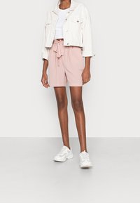 Personne portant une veste blanche courte, un haut blanc court, un short taille haute rose avec une ceinture nouée, et des baskets blanches épaisses.