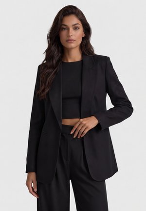 Vrouw met lang donker haar, gekleed in een zwarte blazer, cropped top en bijpassende hoge taillebroek, staand tegen een effen lichte achtergrond.