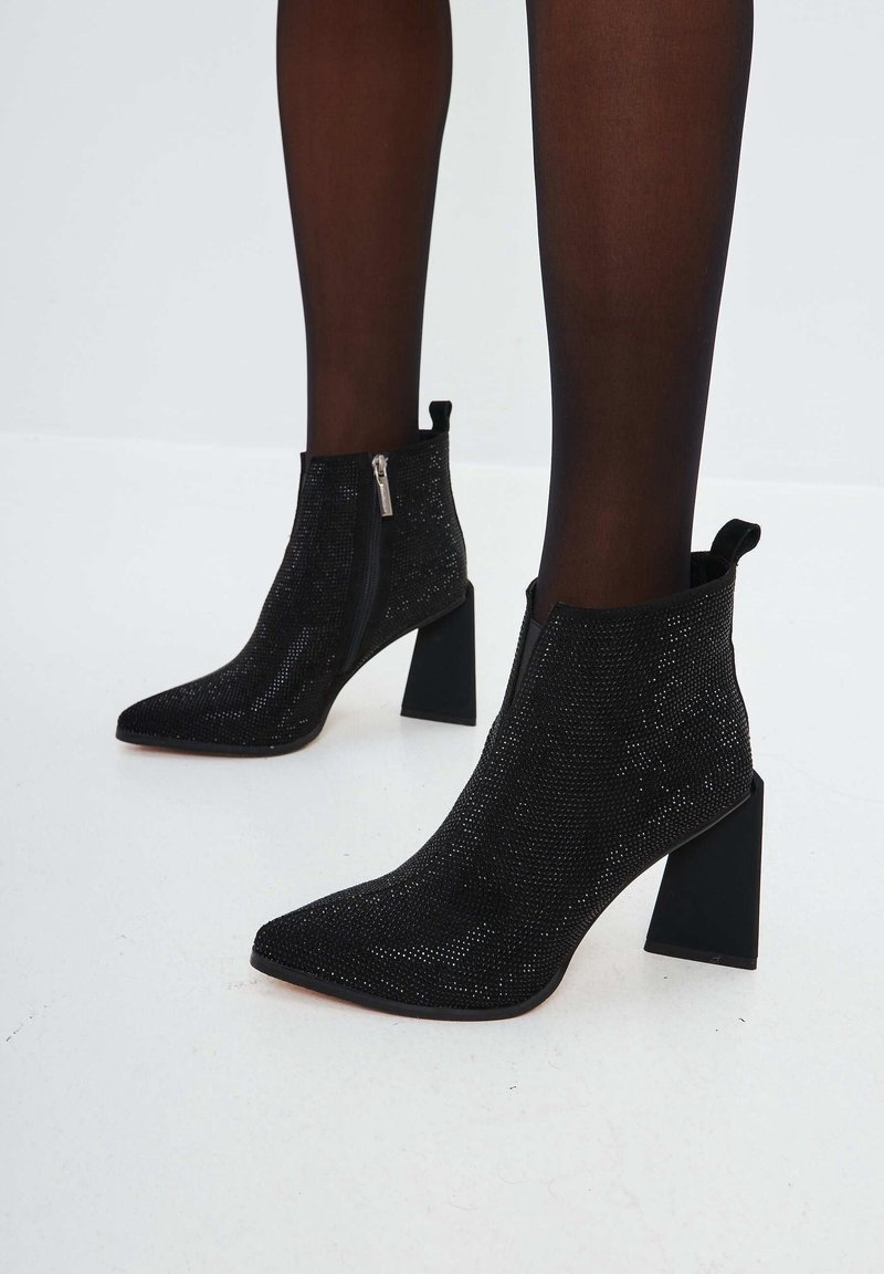Black ankle boots with block heels and side zippers worn with dark sheer tights on a white floor.
