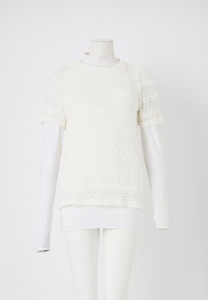 Biely čipkový top s krátkym rukávom a zložitými vzormi, vrstvený nad priliehavou bielou košeľou s dlhým rukávom, vystavený na manekýne.