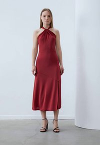 Crvena halter haljina od glatkog materijala, s uskim gornjim dijelom i lepršavom suknjom. Ima bočni prorez i minimalističke crne sandale na remenčiće.