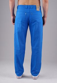 Blaue Baumwollhose mit strukturiertem Finish, ausgestattet mit einem Bund und Gesäßtaschen. Enthält ein Etikett am Bund und weiße Sneakers.