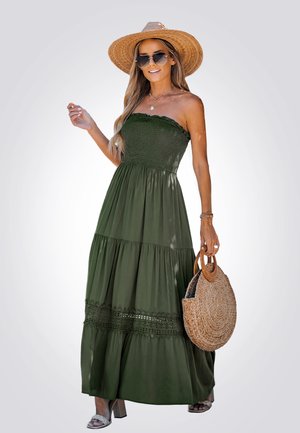 Olijfgroene strapless maxi-jurk met een smock-buste, gelaagde rok en kantaccenten. Gecombineerd met een ronde vlecht tas en sandalen.