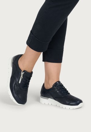 Feet wearing black textured leather sneakers with white soles and side zipper, paired with black rolled-up pants.