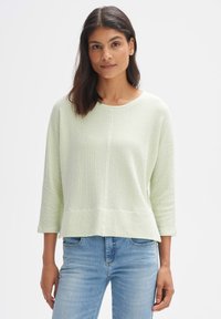 Hellgrüner strukturierter Pullover mit Dreiviertelärmeln und lockerer Passform, kombiniert mit blauen Jeans. Schlichter runder Halsausschnitt und dezente Nähte.