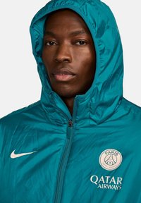 Jachetă cu glugă de culoare teal, cu o textură lucioasă, având închidere cu fermoar, logo-ul Nike și branding-ul Paris Saint-Germain.