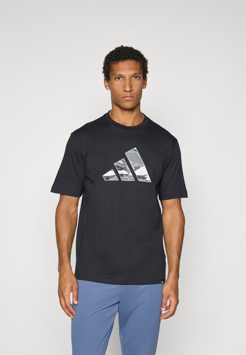 T-shirt en coton noir avec un grand logo Adidas camouflé en blanc et gris sur le devant. Manches courtes et col rond.