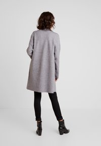 Abrigo gris hasta la rodilla con un corte relajado y textura suave, combinado con pantalones negros y botas de tobillo. Presenta un diseño sencillo y sin adornos.