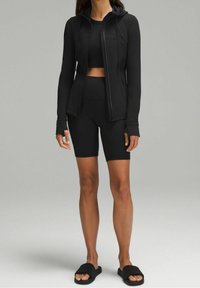 Žena nosící černou mikinu na zip, černý crop top, černé biker šortky a černé sandály na klínku stojí proti šedému pozadí.