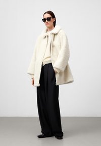 Crème faux fur jas met een brede kraag, oversized pasvorm en textuur oppervlak, gecombineerd met zwarte high-waist broek en zwarte schoenen.