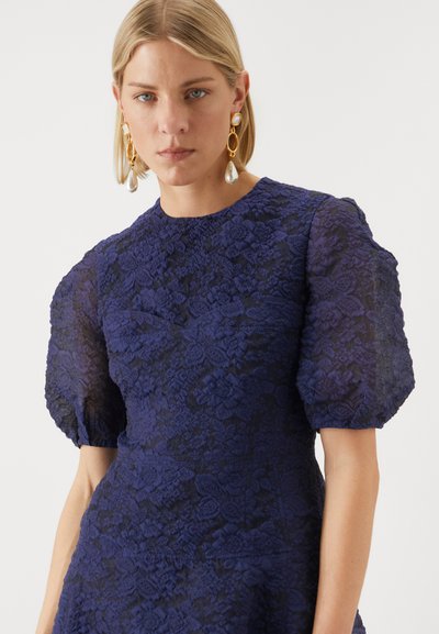 Vestido de renda azul marinho com mangas curtas bufantes, apresentando padrões florais e um acabamento texturizado. Modelo usa brincos dourados pendentes.