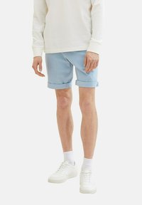Pantalones cortos azul claro con dobladillos enrollados, hechos de tela suave, combinados con una camisa blanca de manga larga y zapatillas blancas, de pie sobre un fondo neutro.