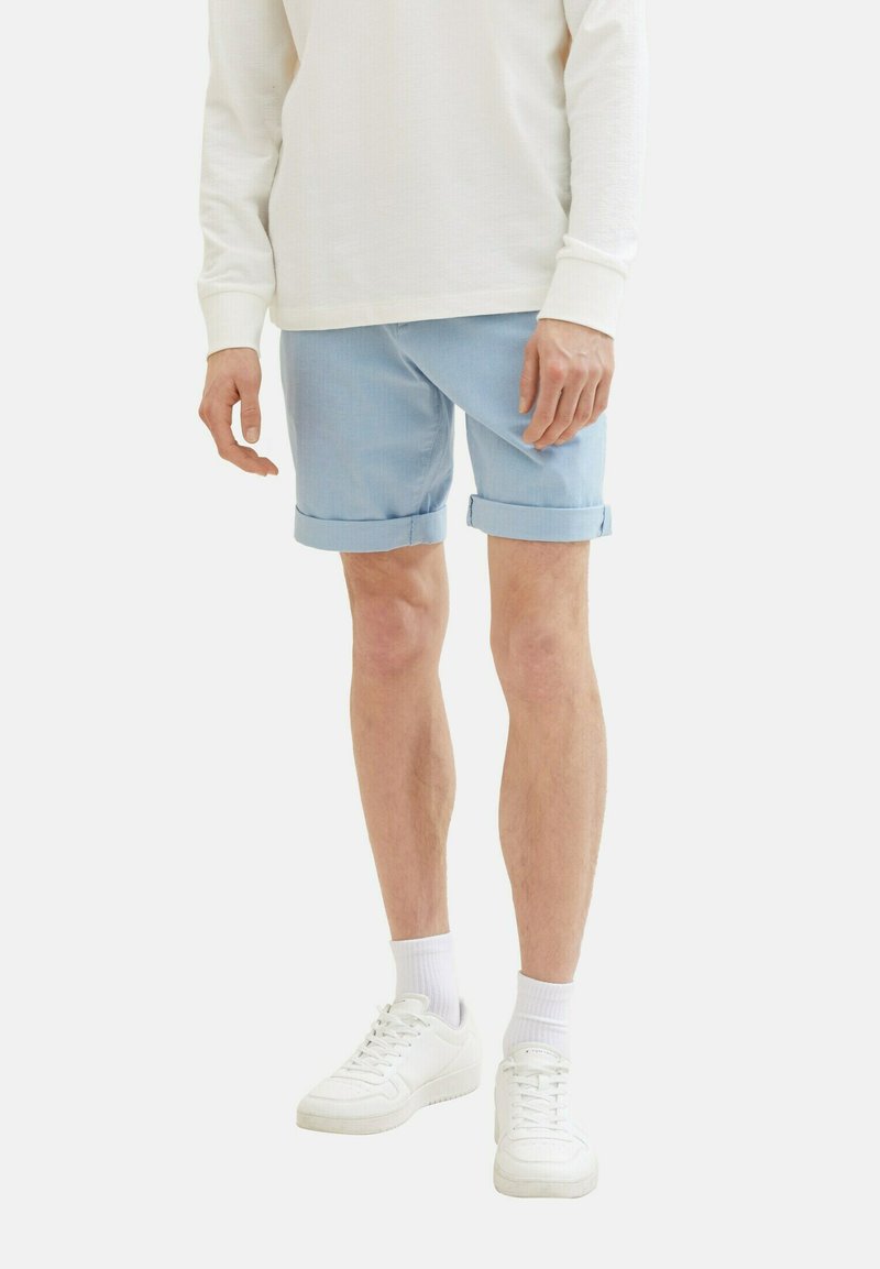 Ljusblå shorts med upprullade fållar, sydda av mjukt tyg, kombinerade med en vit långärmad skjorta och vita sneakers, stående mot en neutral bakgrund.