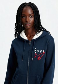 Sweat-shirt zippé navy avec une capuche doublée en sherpa, comportant une broderie "love" en rouge et blanc et un graphique d'épingle de sécurité. Tissu doux et poches doubles.