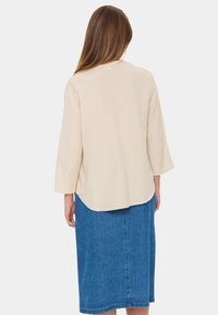 Blusa di lino beige con maniche a tre quarti, orlo arrotondato, abbinata a una gonna di jeans blu. Tessuto morbido, vestibilità casual.