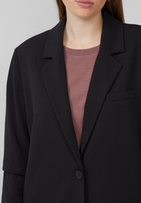 Zwarte blazer met een reverskraag, sluiting met één knoop en een borstzak. Draagt over een mauve shirt, waardoor de gladde stoftextuur zichtbaar is.