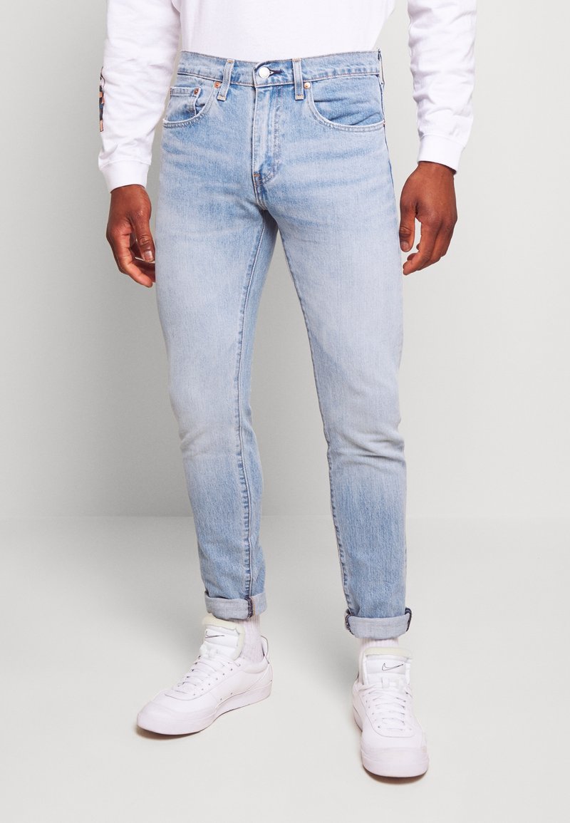 Jean en denim bleu clair, coupe slim, ourlets retroussés, cinq poches et passants pour ceinture, porté avec des baskets blanches et une chemise blanche à manches longues.