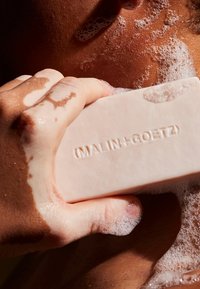 Weißer rechteckiger Seifenblock mit geprägtem Text "(MALIN+GOETZ)", der in einer Hand gehalten wird, umgeben von Blasen auf nasser Haut.