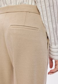 Calças texturizadas em bege com um pequeno bolso traseiro, apresentando um discreto padrão de diamante. O tecido parece respirável e leve.